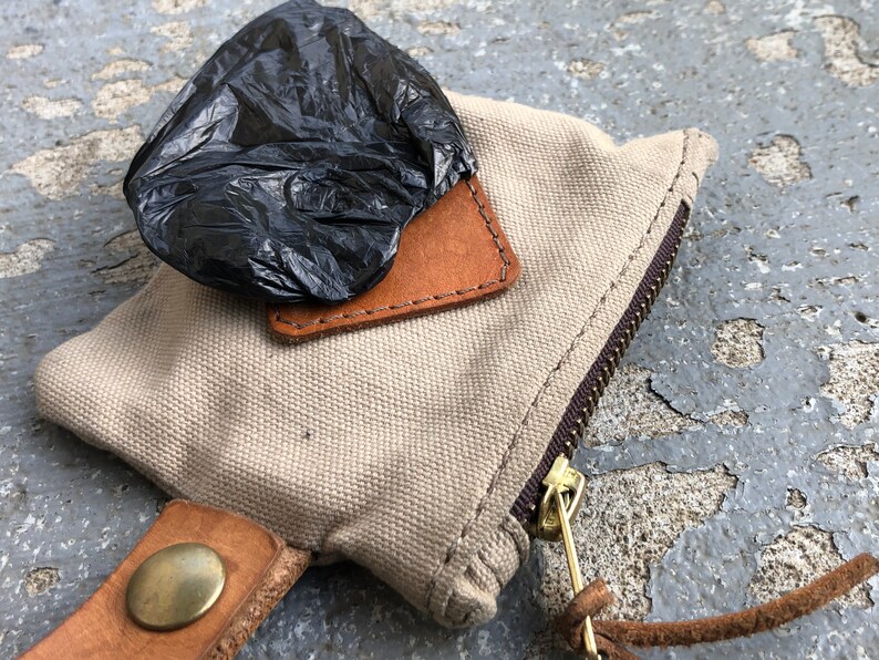 May include: A beige canvas bag with a brown leather flap and a zipper closure. A black plastic bag is on top of the flap. The bag has a brown leather strap with a brass button.