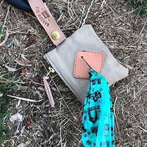 May include: A tan canvas bag with a brown leather flap and a zipper. The bag is attached to a black leather strap with a brown leather tag that says "Ransom Six" and "The Wire". A blue plastic bag with paw prints and the text "The Dog House" is hanging from the bag.