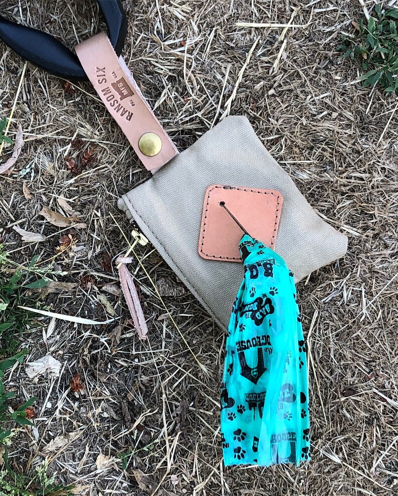 May include: A tan canvas bag with a brown leather flap and a zipper. The bag is attached to a black leather strap with a brown leather tag that says "Ransom Six" and "The Wire". A blue plastic bag with paw prints and the text "The Dog House" is hanging from the bag.