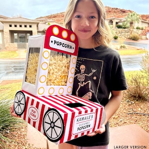 Puede incluir: Una caja de palomitas de maíz a rayas rojas y blancas con las palabras "Popcorn" y "Kamaile's Gourmet Popcorn" impresas. La caja está diseñada para parecerse a un carrito de palomitas de maíz vintage, con ruedas y una ventana transparente.