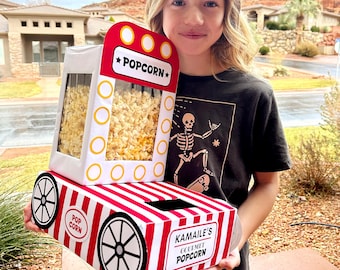 Peut inclure: Une boîte à popcorn à rayures rouges et blanches avec les mots "Popcorn" et "Kamaile's Gourmet Popcorn" imprimés dessus. La boîte est conçue pour ressembler à un chariot à popcorn vintage, avec des roues et une fenêtre transparente.