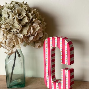 May include: A decorative wooden letter 'C' with a red and white striped design, accented with orange dots. The letter is displayed on a wooden surface. A vase with dried flowers is in the background, with a green glass bottle.