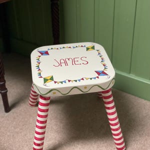 May include: A small wooden stool with a white square seat featuring the name JAMES in red lettering, surrounded by colorful painted kites and bunting. The stool has four cylindrical legs painted with red and white horizontal stripes.