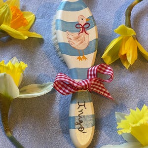 May include: A wooden hairbrush with a light blue and white horizontal striped pattern, featuring a hand-painted light pink duck with a red bow around its neck. The handle is tied with a red and white chequered ribbon and displays the name Inigo.