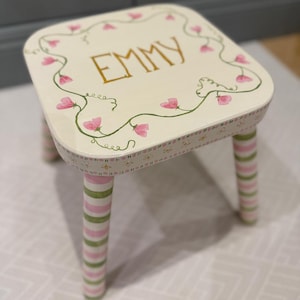 May include: A small, hand-painted wooden stool with the name "EMMY" in gold lettering on the top. The stool features pink floral accents and green vines around the edges. The legs are striped in pink and green.