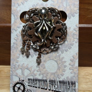 May include: A silver and copper steampunk octopus pin with a green gem in the center. The pin is attached to a gear and has a filigree design. The pin is on a white card with the text "Highwind Steamworks".