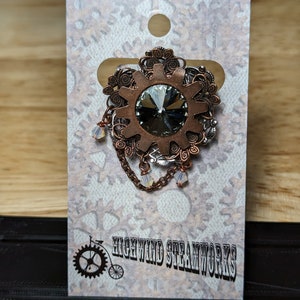 May include: A copper-toned gear with a large, clear crystal in the center, surrounded by intricate filigree. The gear is attached to a chain with small, clear crystals hanging from it. The background is a white card with a black and white illustration of a bicycle with gears and the text "Highwind Steamworks".