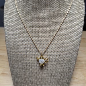 May include: A gold-colored necklace with a teapot pendant. The teapot pendant has a clear, textured center and gold-colored accents. The necklace is displayed on a neutral-colored mannequin.