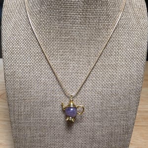 May include: A gold-colored necklace with a teapot pendant. The pendant features a purple gemstone body and gold-colored accents. The necklace is displayed on a neutral-toned mannequin.