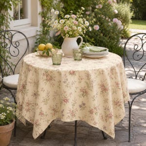 Può includere: Un tavolo rotondo coperto da una tovaglia color crema con un piccolo motivo floreale rosa e verde. Sul tavolo si trovano una brocca in ceramica bianca riempita di fiori bianchi e rosa, una ciotola di limoni gialli, due bicchieri verdi e piatti.