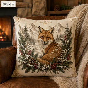 May include: A square throw cushion featuring a printed illustration of a red fox centred against a snowy mountain and pine forest background. The fox is framed by a wreath of green pine branches, pinecones, and red berries with gold accents.