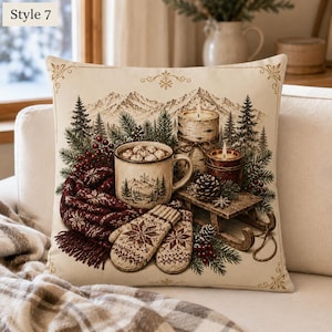 May include: A square throw pillow featuring a printed winter scene with a mug of hot cocoa, a pair of patterned knit mittens, a dark red fringed scarf, two candles, and a small wooden sled, all set against a backdrop of pine trees and mountains.