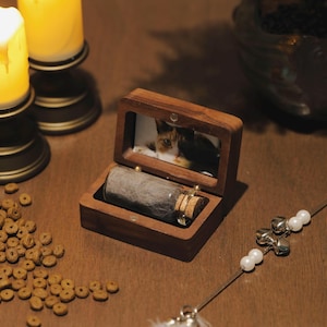 May include: A wooden pet urn with a photo of a cat inside the lid. The urn is open, revealing a small glass bottle filled with pet fur. Cat food and a cat toy are also visible.