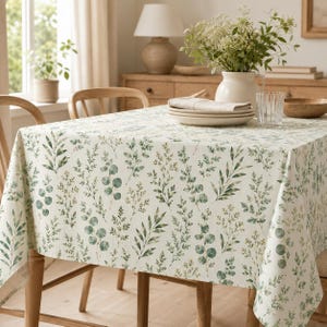 May include: A rectangular tablecloth with a botanical print featuring green leaves and sprigs on a cream background. The tablecloth is draped over a wooden table, with wooden chairs visible. The scene is set in a well-lit dining room.
