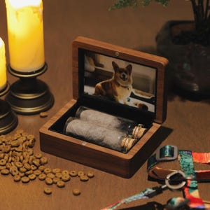 Op de afbeelding: Een houten gedenkbox voor huisdieren met een foto van een hond in het deksel. De doos bevat twee glazen flesjes met kurken die gevuld zijn met dierenhaar. Hondenvoer en een kleurrijke hondenriem zijn ook zichtbaar.