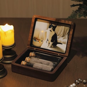 May include: A wooden keepsake box with a framed photo of a black and white cat. The box is open, revealing three glass vials with cork stoppers. The box is on a wooden surface, with a lit candle in the background.