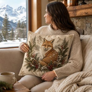 May include: A square throw pillow featuring a detailed illustration of a red fox sitting amidst a winter wreath of pine branches, pinecones, and red berries, set against a backdrop of snow-covered mountains and evergreen trees in muted tones.