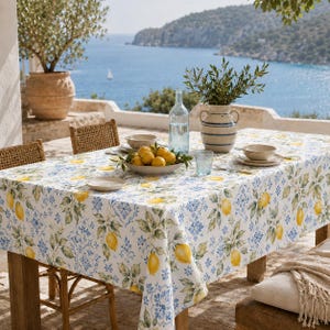 May include: A table set for dining outdoors with a white tablecloth featuring a lemon and floral pattern. The table is set with plates, bowls, and a bowl of lemons. A vase of greenery and a water bottle are also on the table. The background shows a view of the sea.
