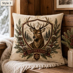 May include: A square throw pillow featuring a central shield graphic with a brown deer head, mountain landscape, and pine trees. The design includes pine branches, pinecones, and red berries, with the text "ALPINE LODGE" on a banner.