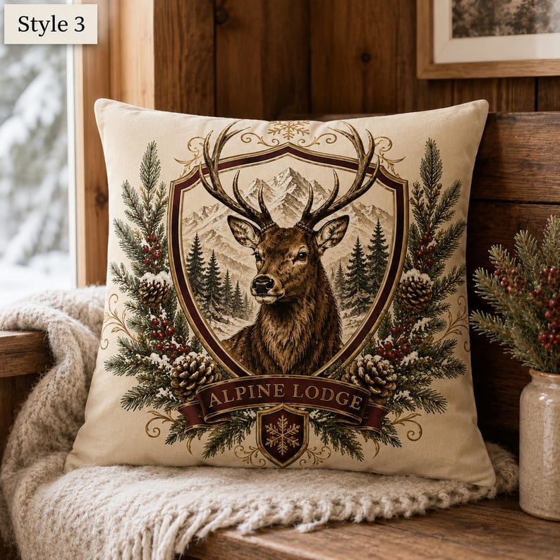 May include: A square throw pillow featuring a central shield graphic with a brown deer head, mountain landscape, and pine trees. The design includes pine branches, pinecones, and red berries, with the text "ALPINE LODGE" on a banner.