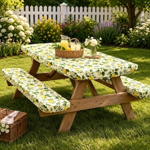 May include: A wooden picnic table with attached benches covered in a white fabric featuring a yellow lemon and green leaf pattern. A woven basket holds a glass pitcher of lemonade and two glasses, with a small vase of white flowers nearby.