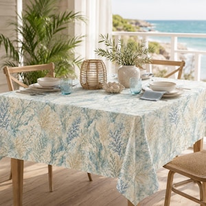 Op de afbeelding: Een eettafel gedekt voor een maaltijd, met een tafelkleed met een blauw en beige koraalrifpatroon. De tafel is gedekt met witte kommen, blauwe glazen en een geweven kandelaar. Een vaas met groen en een decoratief koraalstuk maken het kustthema compleet.