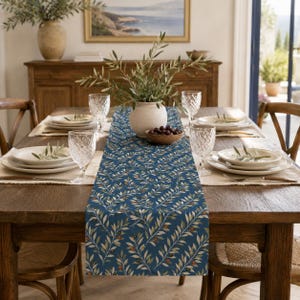 Pode incluir: Um caminho de mesa retangular longo com fundo azul-escuro e um padrão repetitivo de ramos de oliveira verdes, caules finos e pequenas azeitonas vermelhas. O caminho está centrado numa mesa de jantar de madeira com pratos e copos brancos.