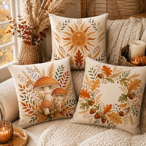 Peut inclure: Trois coussins carrés décorés d'illustrations sur le thème de l'automne. Un coussin présente un visage de soleil central, un autre montre trois champignons à chapeaux tachetés, et le troisième arbore une couronne de feuilles de chêne et de glands.