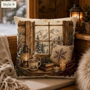 May include: A square throw cushion featuring a printed scene of a window looking out onto snow-covered mountains and pine trees. The design includes a lit lantern, a steaming mug, two stacked books, two pillar candles, and a small snowflake cushion.