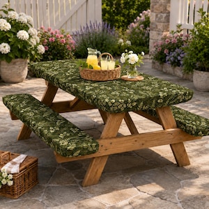 May include: A wooden picnic table set featuring a rectangular table and two attached benches, all covered in a dark green fabric with a white botanical fern and leaf pattern. A woven basket sits on the table holding a glass pitcher and two glasses.