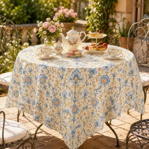 May include: A round table covered with a white tablecloth featuring a blue and orange floral damask pattern. On the table sits a white porcelain teapot, a matching creamer, two teacups with saucers, a two-tier serving stand with pastries, and a vase.