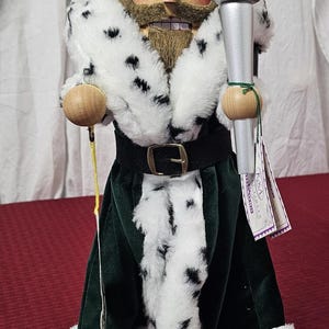 May include: A wooden Christmas nutcracker in the form of a character from "A Christmas Carol." The figure wears a green coat with white fur trim, a black belt, and a fur-trimmed cape. It holds a silver torch with a red and yellow flame.
