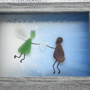 May include: Framed artwork featuring two figures made of sea glass, one green and one brown, holding a white heart. The background is a blue gradient with stars, and the text "Hold on to the LOVE, not the LOSS." is at the top.