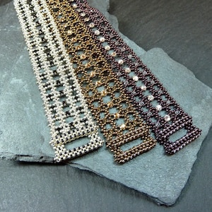 May include: Three beaded bracelets with a geometric pattern. The bracelets are made with silver, gold, and dark purple beads. The bracelets are displayed on a grey stone surface.