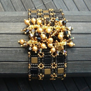 May include: A black and gold beaded bracelet with a geometric pattern and a cluster of gold and black beads hanging from the centre.