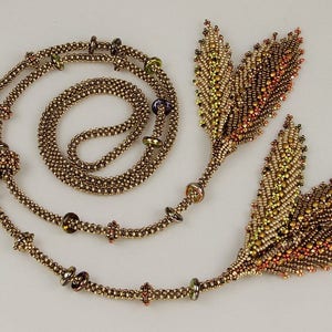 Puede incluir: Un collar de cuentas doradas con dos colgantes en forma de hoja. Las hojas están hechas de pequeñas cuentas en tonos marrones, dorados y naranjas.