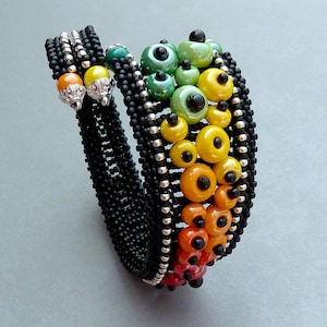 Könnte beinhalten: Ein schwarzes Perlenarmband mit einem mehrfarbigen Muster aus grünen, gelben, orangen und roten Perlen. Die Perlen sind in einem abgestuften Muster angeordnet, wobei die grünen Perlen oben und die roten Perlen unten sind.