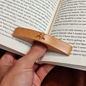 May include: A wooden book page holder with a finger hole, resting on the pages of a book. The holder is light brown with the name "Arlan" written on it. The book is open, with text visible.