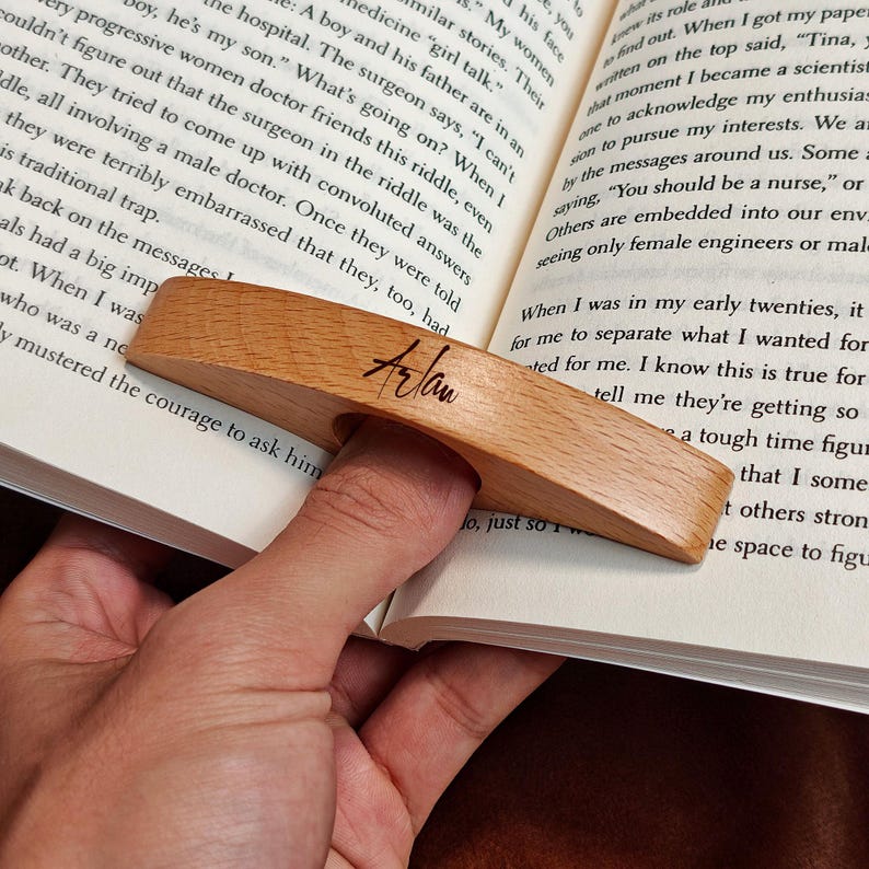 May include: A wooden book page holder with a finger hole, resting on the pages of a book. The holder is light brown with the name "Arlan" written on it. The book is open, with text visible.