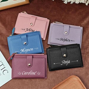 Può includere: Una collezione di portafogli porta carte personalizzati in vari colori: rosa, blu, lilla, bordeaux e nero. Ogni portafoglio presenta un nome stampato in corsivo bianco. I portafogli hanno una chiusura a scatto e una tasca con cerniera.