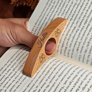 May include: A wooden book page holder with the name "Hollis" engraved on it. The holder has a finger hole and mountain illustrations. The holder is made of light-colored wood and is resting on the pages of a book.