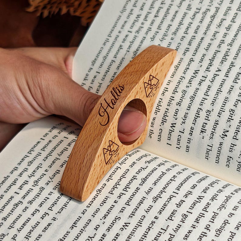 May include: A wooden book page holder with the name "Hollis" engraved on it. The holder has a finger hole and mountain illustrations. The holder is made of light-colored wood and is resting on the pages of a book.