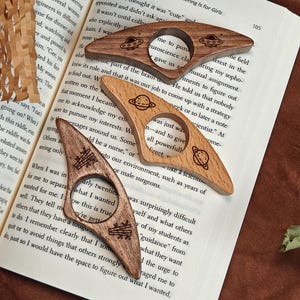 May include: Three wooden page holders with unique designs. One features a planet, another a UFO, and the third has a stack of books. These bookmarks are perfect for book lovers and make a great gift.