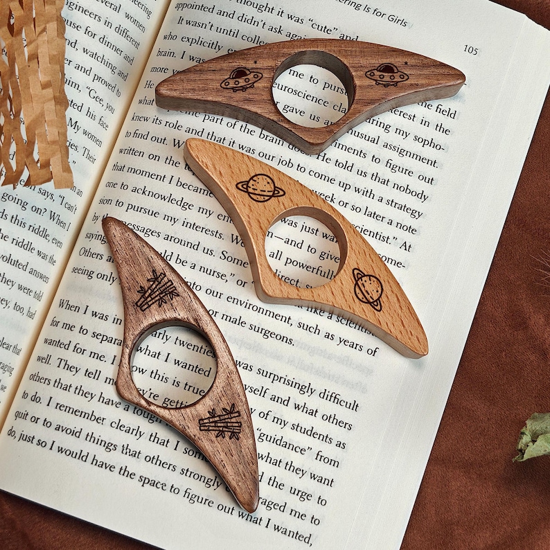 May include: Three wooden page holders with unique designs. One features a planet, another a UFO, and the third has a stack of books. These bookmarks are perfect for book lovers and make a great gift.