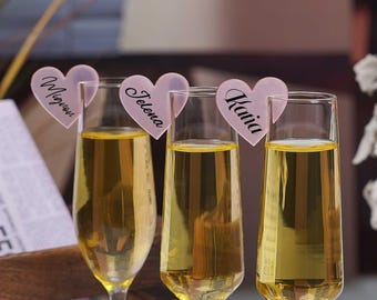 May include: Three champagne flutes filled with a golden liquid, each adorned with a pink heart-shaped name tag. The tags display the names "Mignon", "Jelena", and "Katia" in elegant script, perfect for a celebration.
