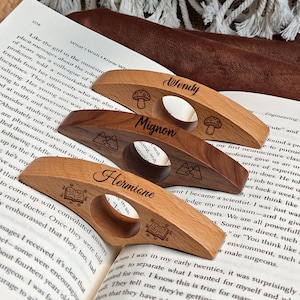 May include: Wooden book page holders with engraved names and decorative icons. The holders are in various shades of brown and are resting on an open book. Each holder has a different name: "Wendy", "Mignon", and "Hermione".