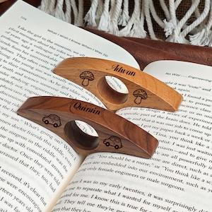 May include: Two wooden book page holders, one light and one dark, rest on an open book. The light-colored holder has the name "Adwan" and mushroom designs. The dark-colored holder has the name "Quintin" and car designs.