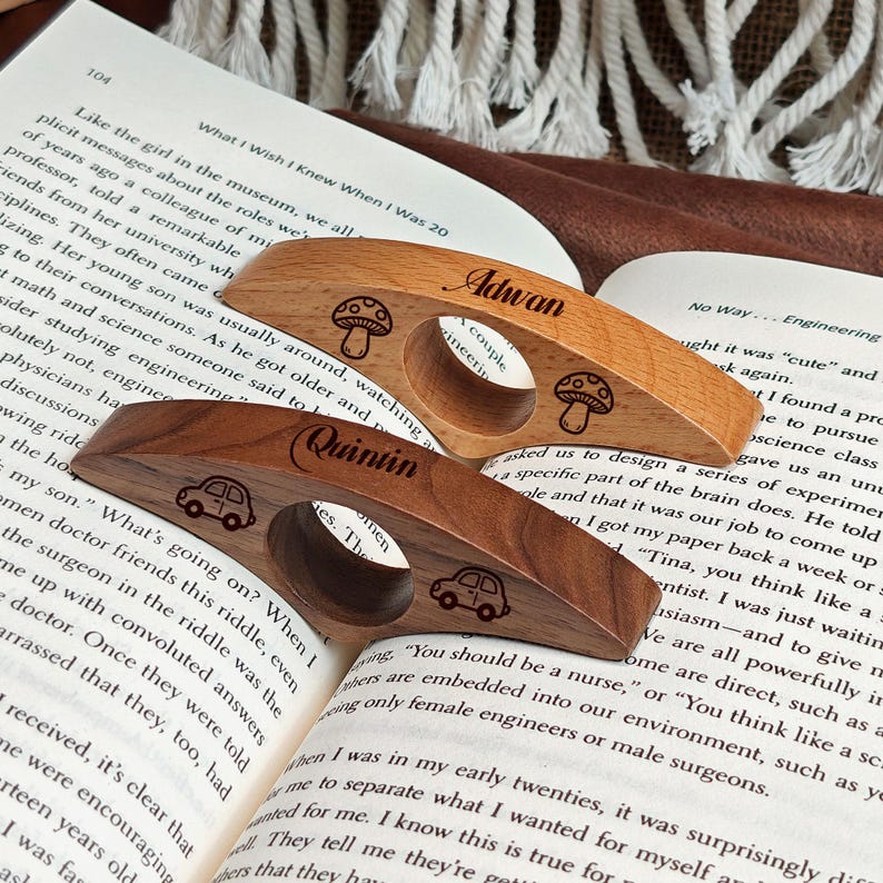 May include: Two wooden book page holders, one light and one dark, rest on an open book. The light-colored holder has the name "Adwan" and mushroom designs. The dark-colored holder has the name "Quintin" and car designs.