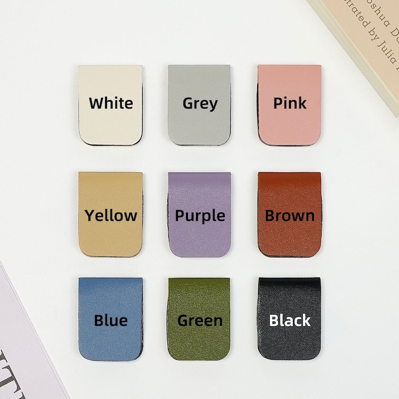 May include: Nine leather bookmark tabs in different colors: white, grey, pink, yellow, purple, brown, blue, green, and black. Each tab has the color name printed in black.