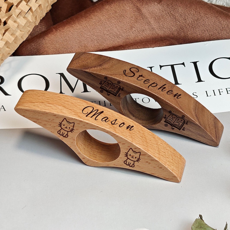 May include: Two wooden thumb page holders. One is light-colored with a cat design and the name "Mason." The other is dark-colored with the name "Stephen" and a book design. Both have a circular opening.